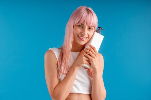 Happy woman with smooth pink hair holding and hugging pump bottle with hair mask or shampoo, posing isolated over blue studio background. Beauty and hair care concept