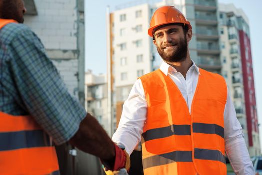 Two men engineers in workwear shaking hands against construction site, close up