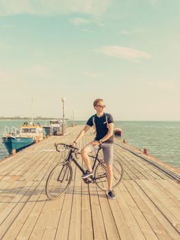 Handsome guy standing with urban bicycle on wooden pier on background of sea in summer. Toned image.
