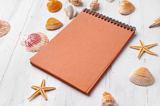Brown open notepad surrounded by sea shells on white wooden table, copy space