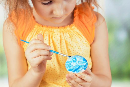 Cute little girl paints egg in blue color for Easter day indoors
