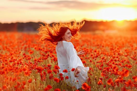 Happy redhead smiling woman in white dress on field of poppies at warm summer sunset