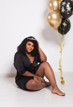 Portrait of smiling young African-American adult woman looking sweet on yellow background holding balloons.