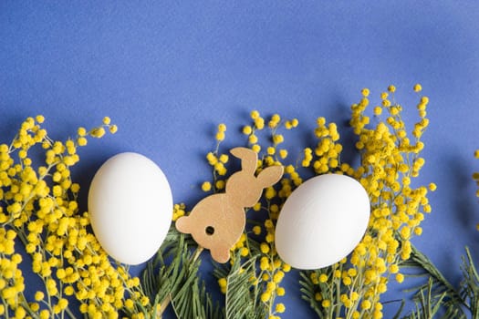 Easter background, white eggs and a wooden hare on a blue background, decorated with Mimosa flowers, flatlay, top view, empty space for text. happy Easter.
