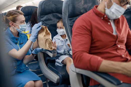 Cute girl in protective face mask looking at female flight attendant serving lunch to little passenger on board. Traveling by airplane during Covid19 pandemic