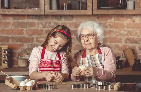 Elderly woman teaching a little girl in red apron to bake homemade cookies spreading dough with rolling pin