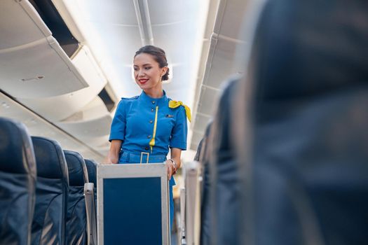 Smiling female cabin attendant leading trolley cart through empty plane aisle. Travel, service, transportation, airplane concept