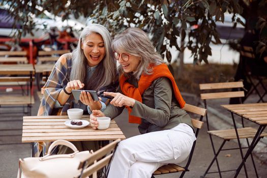 Positive senior Asian lady with friend surf internet on smartphone sitting at small table in street cafe on nice autumn day