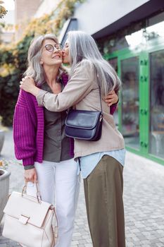 Mature Asian woman hugs and kisses positive female friend with glasses meeting on modern city street. Long-time friendship relationship