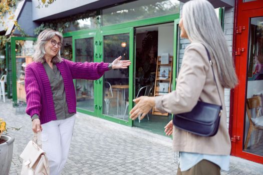 Happy senior woman with spectacles meets silver haired best friend walking along modern city street. Long-time friendship relationship