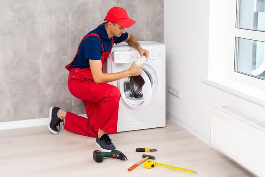 Plumber in overalls with tools is repairing a washing machine in the house