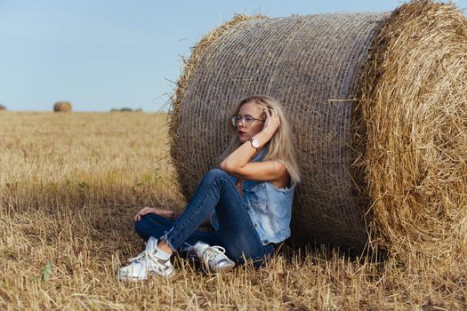 Beautiful girl villager posing in jeans near a bale of hay in a field