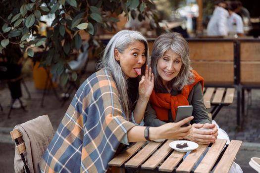 Mature Asian woman grimaces and silver haired friend at videochat via smartphone sitting at small table in street cafe on nice autumn day