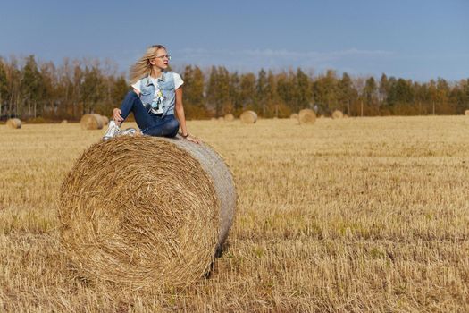 Beautiful girl villager posing in jeans on a bale of hay in a field