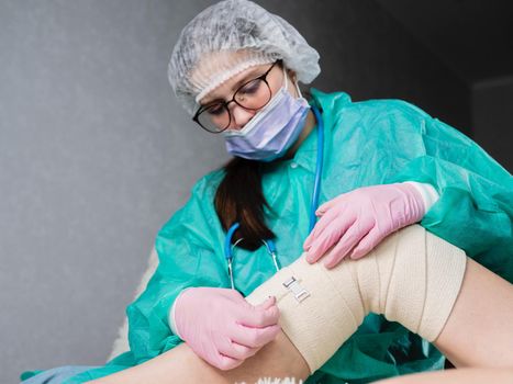A doctor wearing medical gloves wraps an elastic bandage around the patient's leg and knee. The traumatologist applies an elastic material to the patient