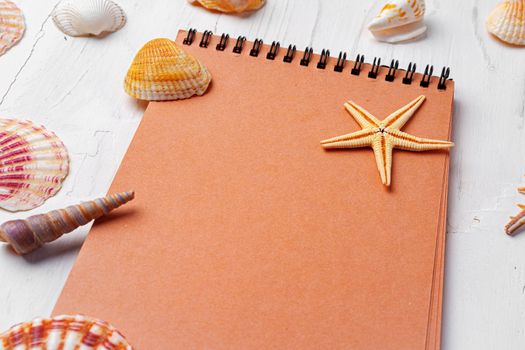 Brown open notepad surrounded by sea shells on white wooden table, copy space