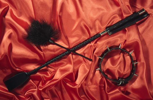 Bondage, kinky adult sex games, kink and BDSM lifestyle concept with a whip, feather stick, collar on red silk with copy space