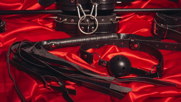 Bondage, kinky adult sex games, kink and BDSM lifestyle concept with a pair of leather handcuffs, flogger, ball gag and a coller with a leash attached on red silk with copy space