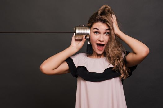 portrait of young surprised woman hearing through a tin can over grey.