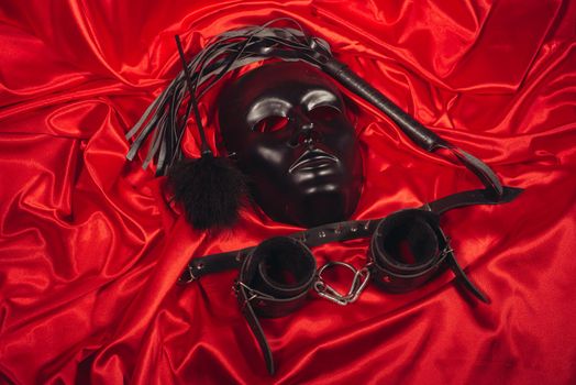 Bondage, kinky adult sex games, kink and BDSM lifestyle concept with a mask, pair of leather handcuffs, flogger, ball gag and a coller with a leash attached on red silk with copy space