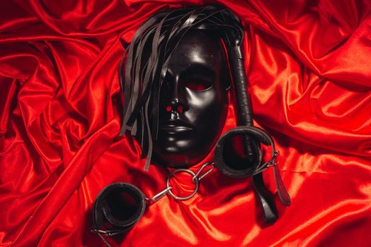 Bondage, kinky adult sex games, kink and BDSM lifestyle concept with a mask, pair of leather handcuffs, flogger, ball gag and a coller with a leash attached on red silk with copy space