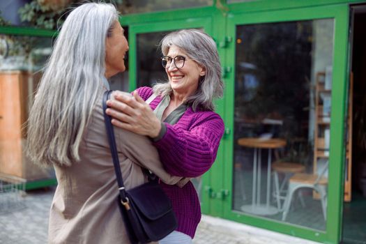 Smiling mature woman embraces female friend with long silver hair meeting on modern city street. Long-time friendship relationship