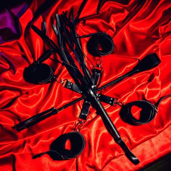 Top view of bdsm outfit. Bondage and whip crossed spank on the red linen. Adult sex games. Kinky lifestyle. - image toned