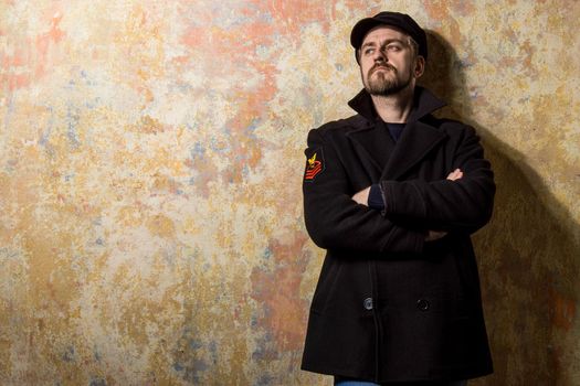 attractive young bearded man in a pea coat and cap is standing at the wall