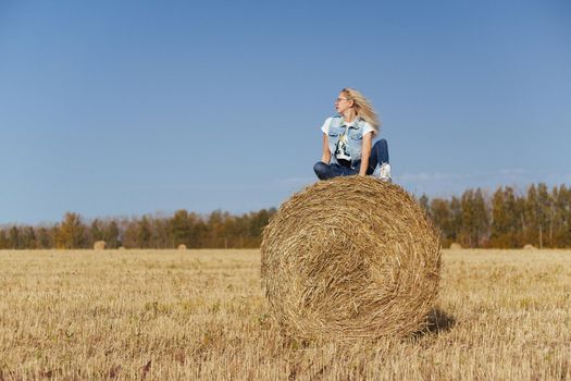 Beautiful girl villager posing in jeans on a bale of hay in a field