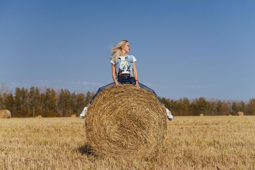Beautiful girl villager posing in jeans on a bale of hay in a field