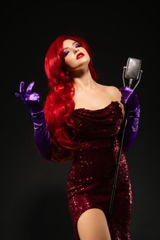 Young romantic redhead woman with very long hair in red dress with microphone on the stand sings with his eyes closed on a black background. Sexy gown on a beautiful slim figure. Long pink gloves