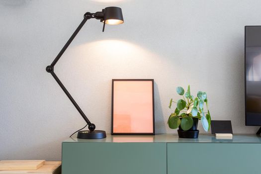 Stylish modern interior, empty picture frame with black lamp and green Chinese Money plant, retro pancake plant on green table scandinavian design close-up