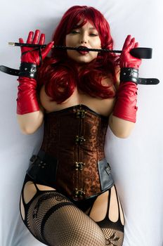 Red-haired woman in a red wig, corset and leather gloves posing lying on a bed holding a stick in the hands of a whip. Bdsm outfit