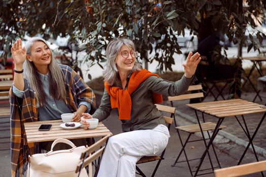 Pair of mature women friends with long grey hair wave hand calling waiter at small wooden table in street cafe on nice autumn day