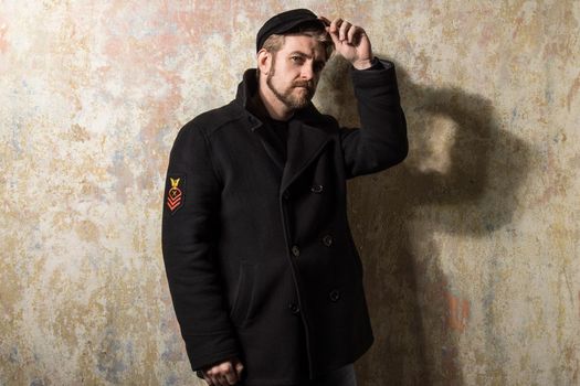 attractive young bearded man in a pea coat and cap is standing at the wall