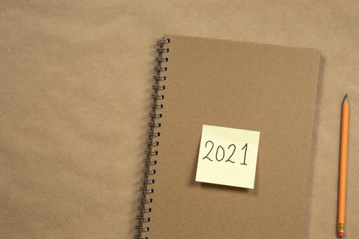 Top view of Brown notepad and yellow sticky note with 2021 New Year's message, pencil on brown paper background texture, education or business concept copy space