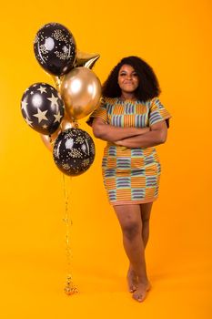 Portrait of smiling young African-American adult woman looking sweet on yellow background holding balloons.