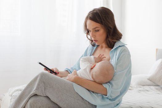 Young mother breastfeeding her child and browsing smartphone on bed.