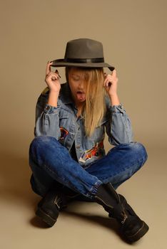 Teenage girl sitting on the floor wearing a hat, jeans, jacket, shoes and showing the tongue