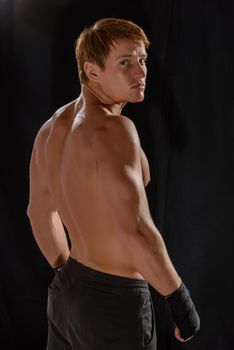 Rear view of strong young male boxer. Fitness male model wearing boxing gloves standing on dark background.