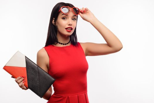 fashionable surprised young lady in a red dress and clutch holds on to sunglasses in the shape of a heart.