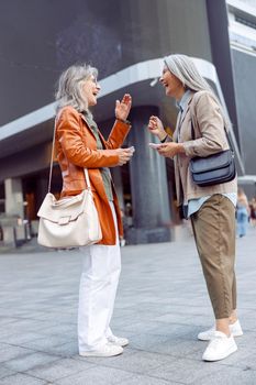 Joyful silver haired senior women with modern mobile phones laugh standing on city street on nice autumn day. Old friends meeting
