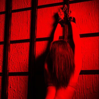 BDSM outfit for adult sex games. A young woman in red light chained to the bars with handcuffs with bondage awaiting punishment.