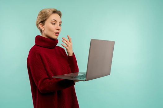 Surprised girl in a red sweater looks at a laptop screen on a blue background - image