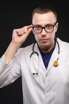 Portrait of a young Caucasian doctor in glasses and a white coat. Holds glasses and looks slyly at the camera.