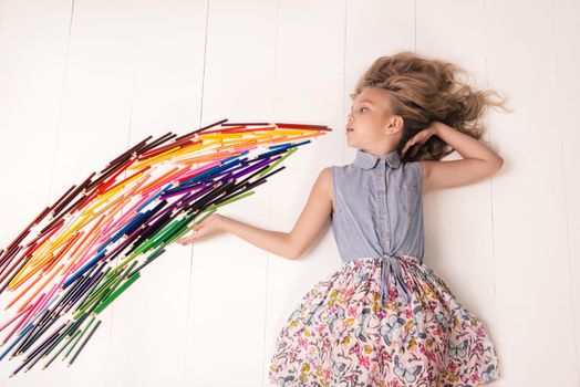 A nice young girl in a dress with butterflies lies on the floor and blows a stream of colored pencils