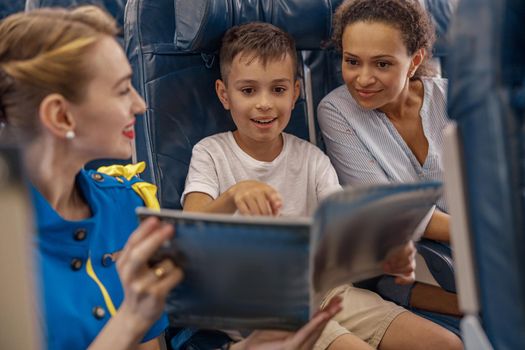 Female flight attendant entertaining a kid on board by offering a book to read. Cabin crew provide service to family in airplane. Airline transportation and tourism concept