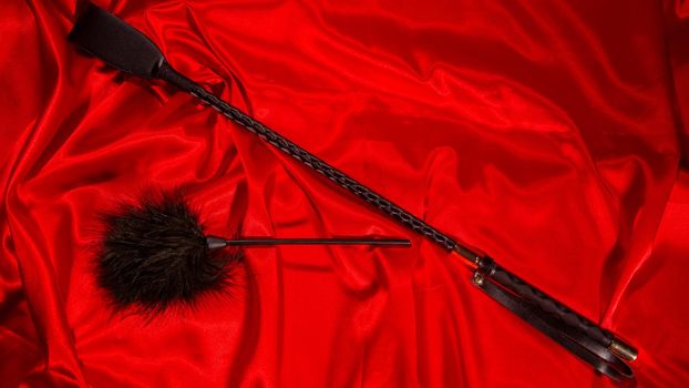 Bondage, kinky adult sex games, kink and BDSM lifestyle concept with a whip, feather stick on red silk with copy space