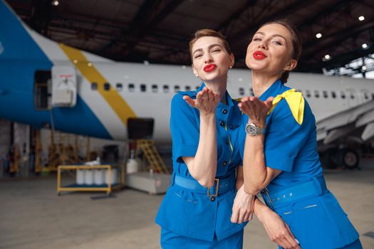 Two air stewardesses in stylish blue uniform blowing kisses to camera, standing together in front of passenger aircraft in hangar at the airport. Occupation concept