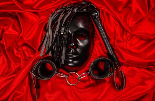 Bondage, kinky adult sex games, kink and BDSM lifestyle concept with a mask, pair of leather handcuffs, flogger, ball gag and a coller with a leash attached on red silk with copy space